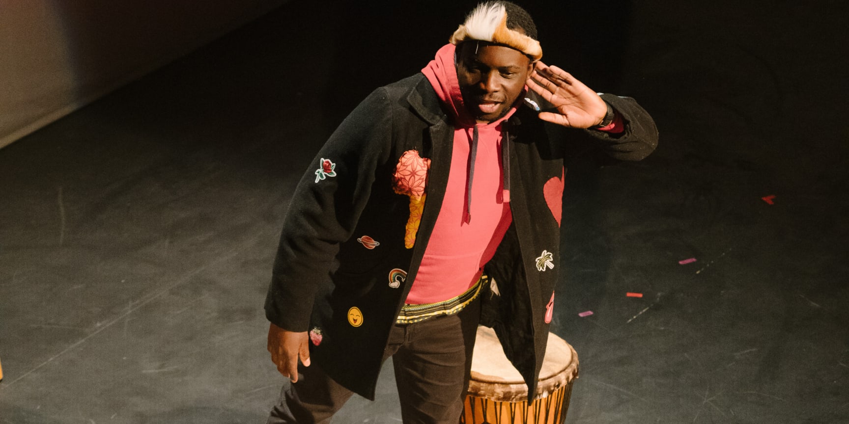 Butshilo Nleya, a  black man wearing black jeans, a black jacket, a pink top and trainers stands on a stage next to a drum with his hand to his ear as if listening for the audience.