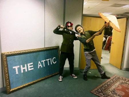 A photo of two performers holding an umbrella and batton, next to sign that says The Attic in an indoor space
