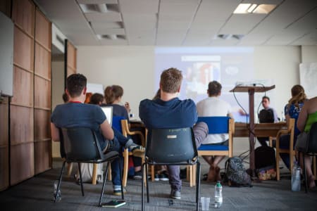 A photo of people sat on chairs watching a presentation from a projector with laptops and notepads, in a room with bright lighting