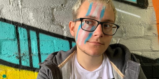 Link to Self-Made Man page. Cas, a transgender man with partially bleached hair and black rimmed glasses, has the trans flag painted on his face, smiles slightly and looks slightly to the left.