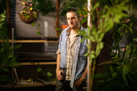 A photo of a person standing in the middle of green plants in an indoor space, the person is wearing a checked top and has one hand resting on their jeans