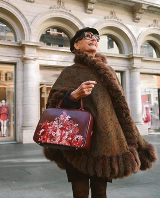 A woman standing in a bright city street, modelling a red bag. She is wearing a brown fur trimmed coat, black frame glasses and a black hat. She is looking smiling up at the sky. The reg bag hangs off her arms and is covered in various sahde of red s