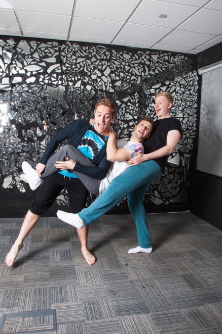 A photo of three people posing in front of an abstract dark wall indoors, the room is light with striped carpet tiles on the floor