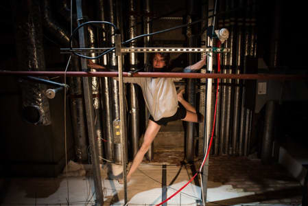 A photo of a person posed between metal bars and wires in a dark space with theatrical lighting, they are wearing a white top and black shorts