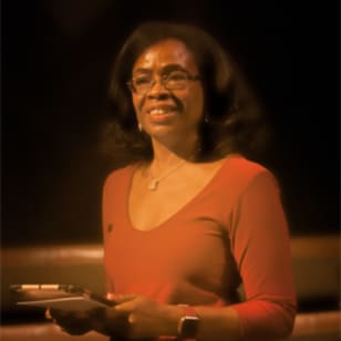 Pam Fraser Solomon, a Black woman with mid-length black hair, wearing an orange top. She smiles and  holds a paper like she is doing a speech.