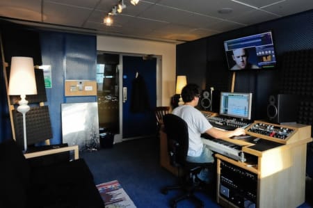 A photo of a person working at a technical desk with a screen above him in a room with blue walls