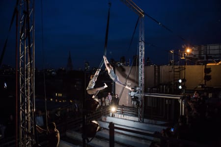 A photo of two people doing acrobatics from large scaffolding outside with audience members watching below, the photo is dark with theatrical lighting