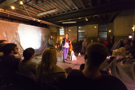 A photo of a person performing in an industrial-looking space to an audience sitting around them