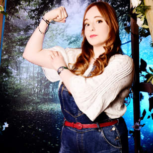 Hannah, a white woman with long red hair wears dungarees and a white tshirt. She stands flexing her bicep.
