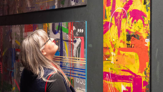 A woman with glasses and long grey hair looks up at abstract paintings on a wall