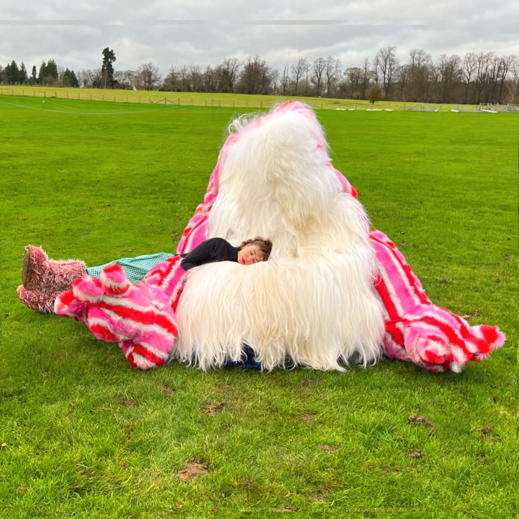 A green field, a young woman rests her head on what looks like a giant, colourful, furry sofa cushion monster