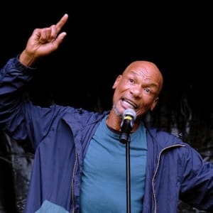 Errol McGlashan, a Black man, wears a blue t-shirt under a dark blue bomber jacker and points his finger in the air mid speech 
