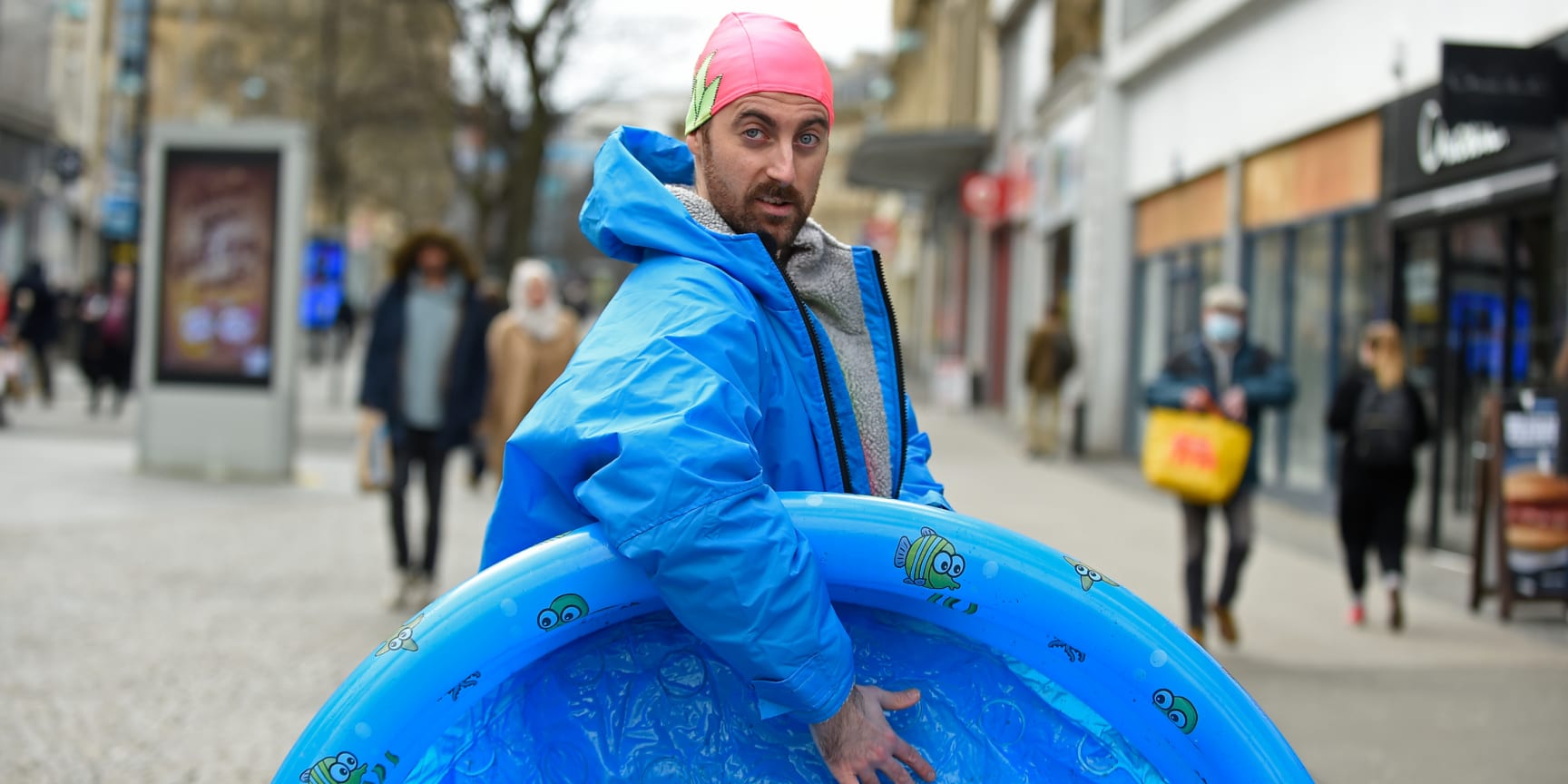 Charlie Hammond, a white man in silly blue waterproofs and a pink swimming cap holds a blue paddling pool in a city centre and looks faux seriously at the camera.