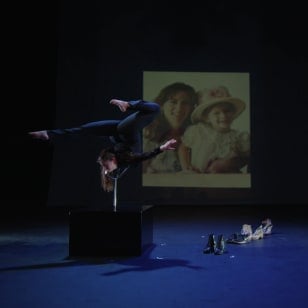 On a dimly lit stage, a contortionist bends on a stand with her arm outstretched in front of a project.