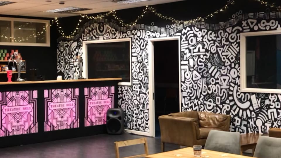 Theatre Delis bar area with a monochrome mural, pink back lit art deco panels on the bar and fairy lights on the wall. In the foreground is a table with a candle in the centre and chairs around it.