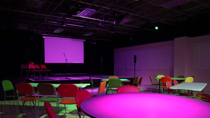 Theatre Delis space set up for a conference with round and rectangular tables set up with chairs in front of a stage with a screen and microphone. The lighting is pink and green, accentuating the chairs laid out.