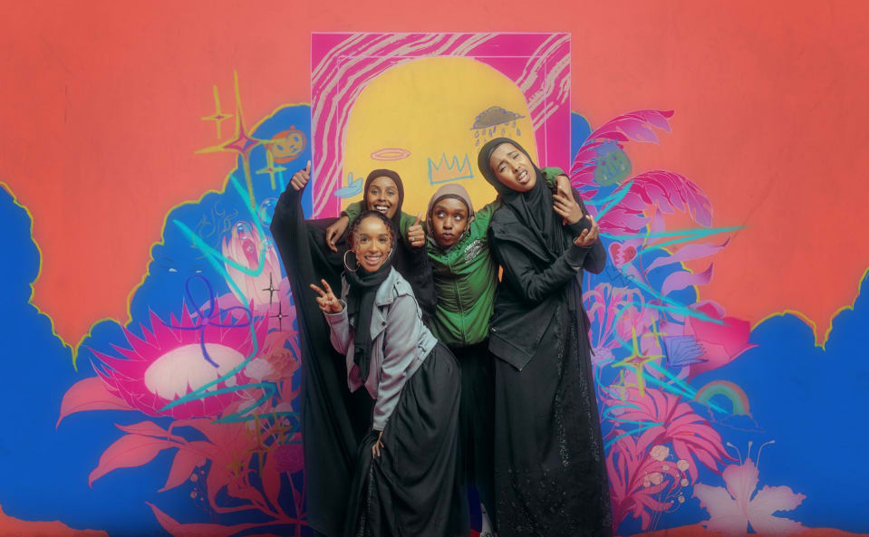 Four young, Black Somali girls are posing in front of a colourful mural, which has been drawn over with youthful, exuberant graffiti.