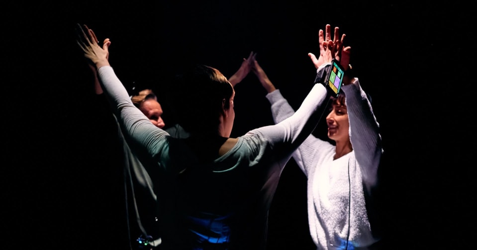 A group of 3 people in a dark environment forming a circle and connecting with their arms towards each other and their hands touching at the fingertips. Their body is angled and is in movement. Two of them, the audience members, are wearing headphone