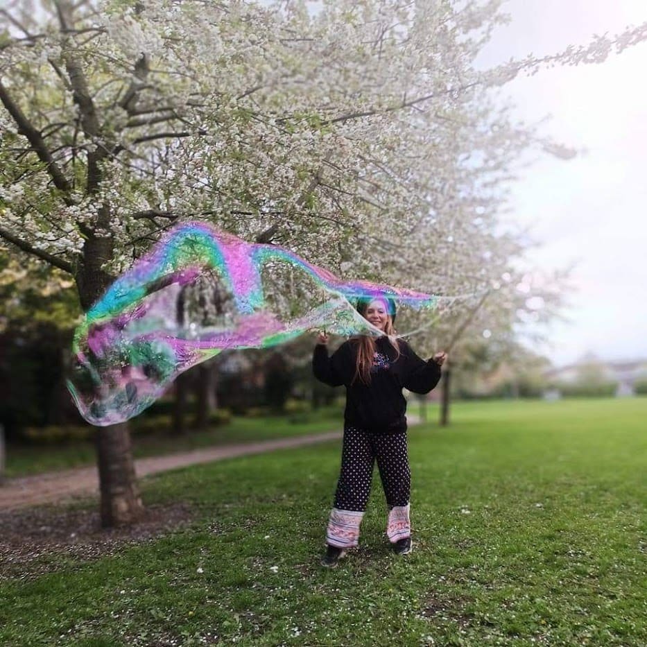 A woman is standing in a park creating large bubbles. She is a white woman who is wearing polka dot trousers, a black jumper and a black hat