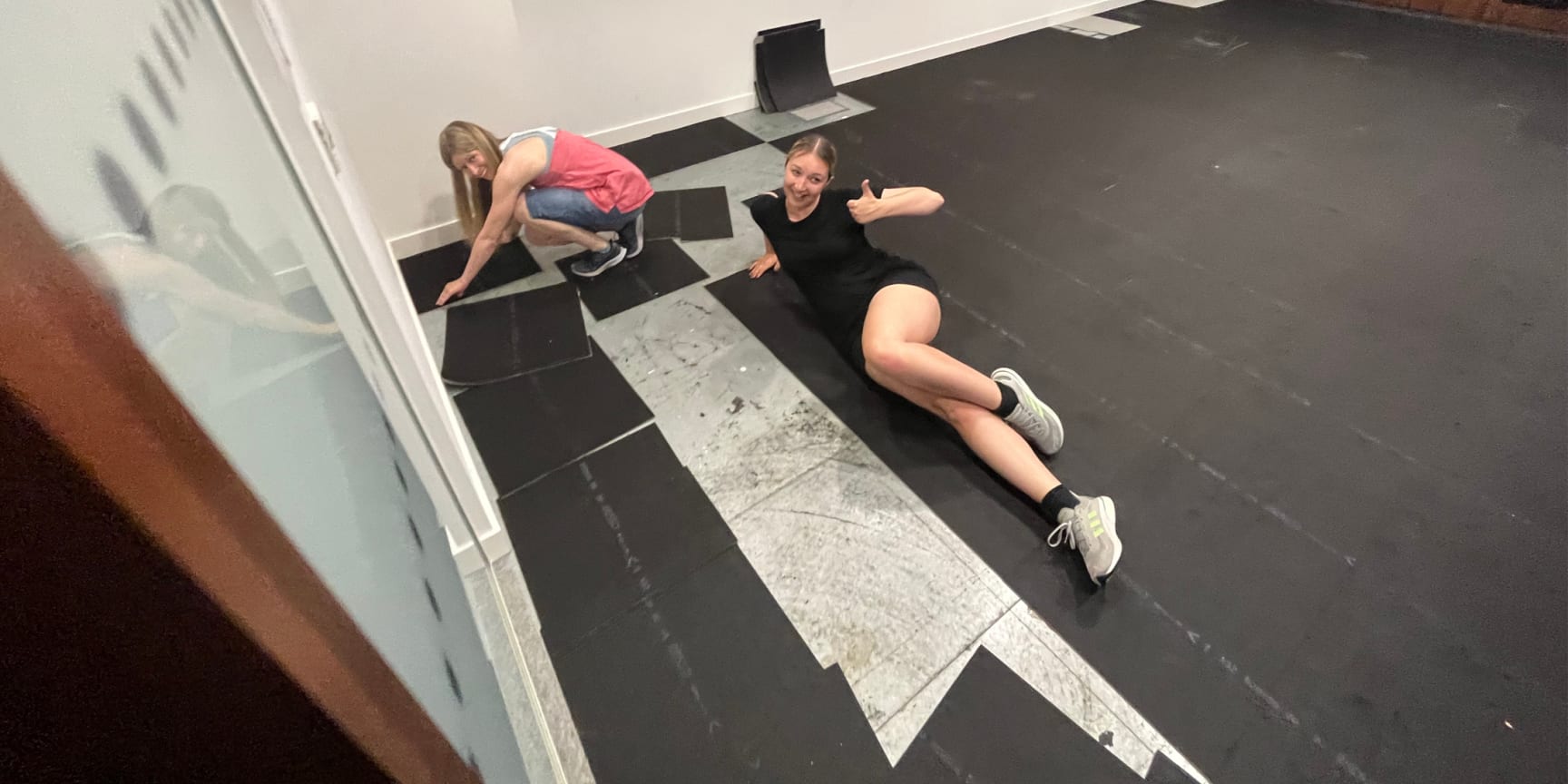 Two white women smile at the camera and lay down dance floor tiles. One poses with her leg in the air.