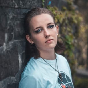 Isobel, a white woman with brown hair leans against a stone wall wearing a t-shirt and blue eye shadow.