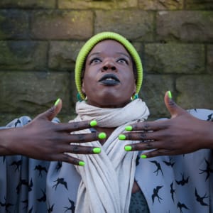 Naomi, a Black person looks up to the sky wearing a bright green woolly hat and holds her matching green nails in front of her scarf.