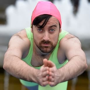 A white man in a swimming hat and tank top poses in a diving position facing the camera in front of a fountain.