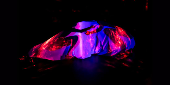 Link to wake up octopus. On a dark stage is a bubble of blue fabric lit from the inside by purple light and red dappled projections, wrapped around two crouching figures. They face one another, only discernible as silhouettes, reaching their arms toward each other, but never quite touching. Photo by Lidia Crisafulli. 