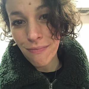 An image of a person’s face, they are white with dark brown short curly hair and dark brown eyes, smiling, wearing a fuzzy dark green jacket.