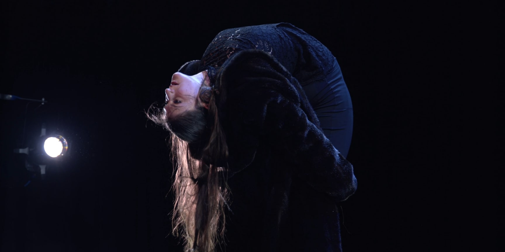 Hannah Finn, a white woman wit long brown hair, bends backwards on a dimly lit stage