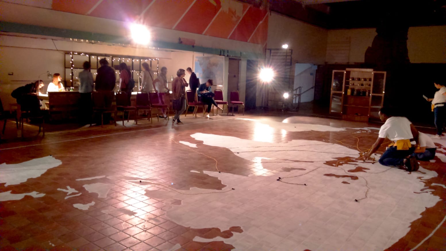 A photo of a large white map drawn onto a tiled floor. A person adding details to the map and people standing around the room