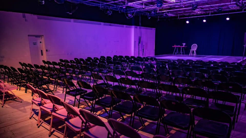 Theatre Delis performance space lit in purple with black fold out chairs set up facing the stage