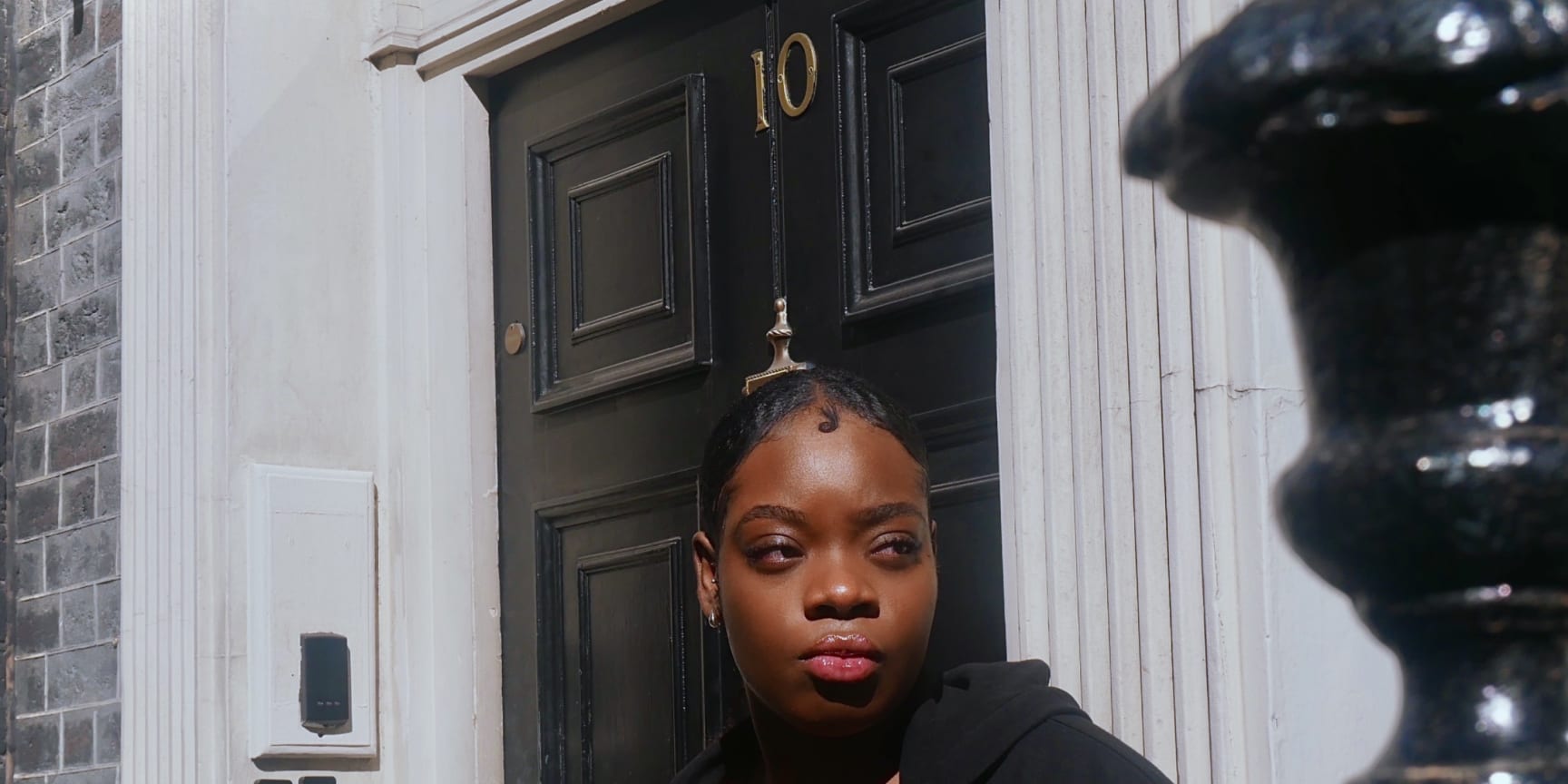 Zakiyyah, a Black woman in hear early twenties, leans against the railing outside no. 10 Downing Street