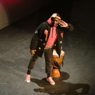 A black man wearing black jeans, a black jacket, a pink top and trainers stands on a stage next to a drum with his hand to his ear as if listening for the audience.