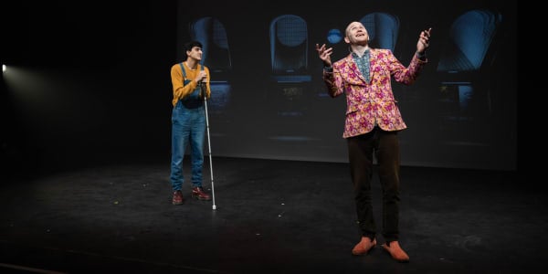 Aarian and Sam from FlawBored stand on stage. Aarian wears dungarees and holds a  cane while Sam wears a jazzy suit and looks animated.
