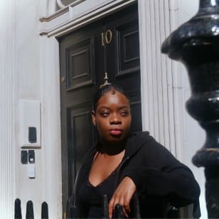 Zakiyyah, a Black woman in hear early twenties, leans against the railing outside no. 10 Downing Street