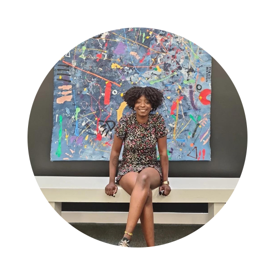 Nicole-Rose, a Black woman with an afro wearing a flowery dress and Converse shoes, smiles and sits on a bench in front of her artwork at Theatre Deli