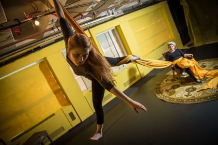 A photo of a person with long hair doing acrobatics from the ceiling and a person sitting on a chair on a rug, they are in front of a yellow wall in an indoor performance space