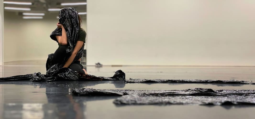 A white woman sitting on the floor with black packaging film wrapped around her body, covering her face. She is resting her chin on her palm.