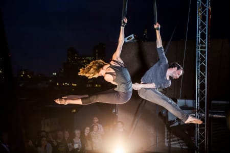 A photo of two people hanging from scaffolding outside and holding hands, it is dark with theatrical lighting and people are watching from below