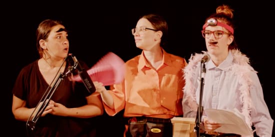 Link to Radiophonics. Three women stand on stage wearing comical outfits - one with a moustache on their head looking suprised, another looking serious with a fake moustache and a feather bower
