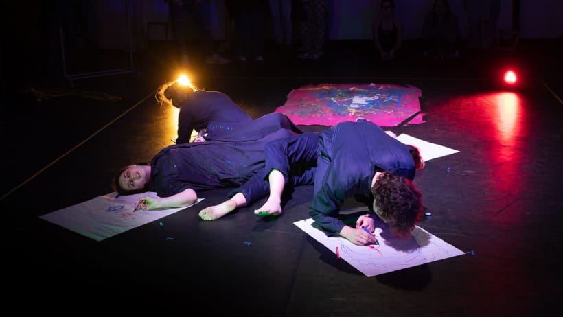 Three people in boiler suits sprawled out on the floor, they are drawing on pieces of large paper and covered in paint