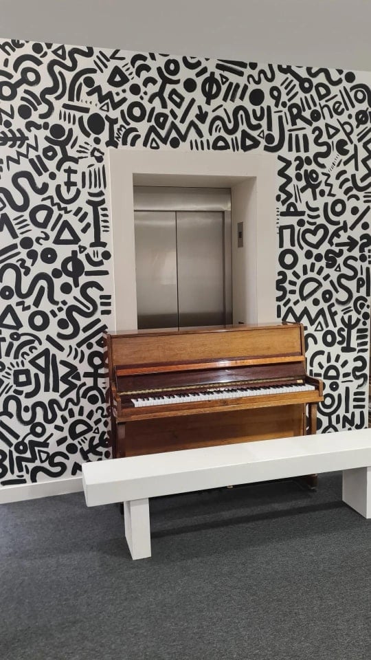 A piano sits infront of a lift, the wall of which is covered in fun abstract designs by Nicole-Rose