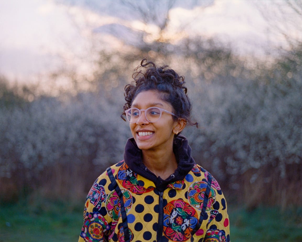 An image of Gayanthiri in a field, they are looking off to the right smiling. They are wearing glasses. they have their hair in a bun on top of their head and they are wearing a bright patterned yellow jacket
